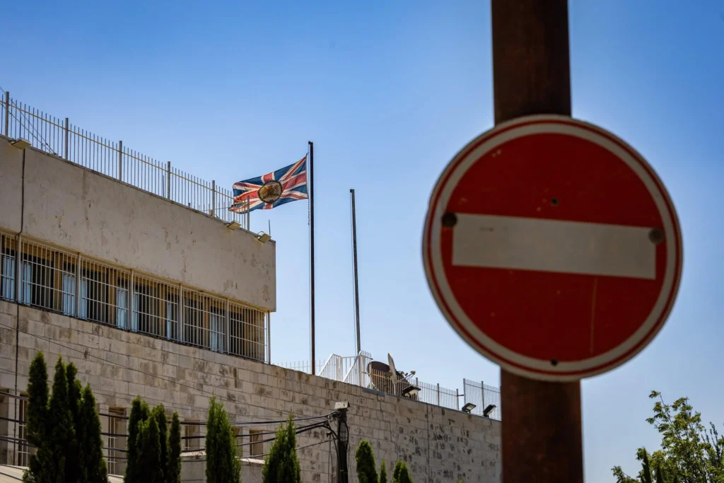 A photograph of a stone British embassy building with a Union Jack flag flying above it, partially framed by a prominent red "No Entry" sign in the foreground, symbolizing the diplomatic chill and growing hostility that casts a dark shadow over British Jews as Britain targets Israel.