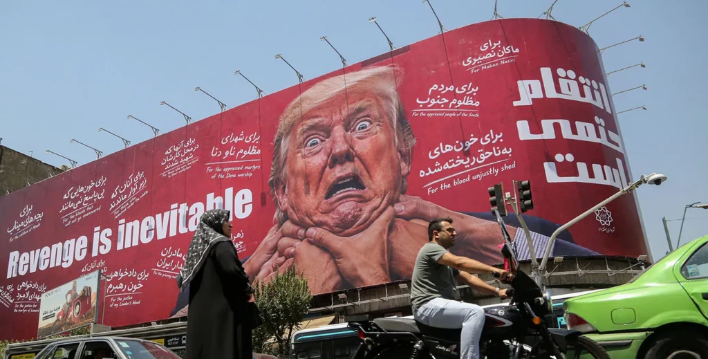 Three Ruptures Are Ending Pax Americana in the Middle East A large red billboard in an Iranian street displays a distressed image of Donald Trump with hands gripping his neck and the slogan "Revenge is inevitable," along with Persian text, as a woman in a black chador and a motorcyclist pass beneath it — a visual sign of the anti-American sentiment that signals the ruptures ending Pax Americana in the Middle East.