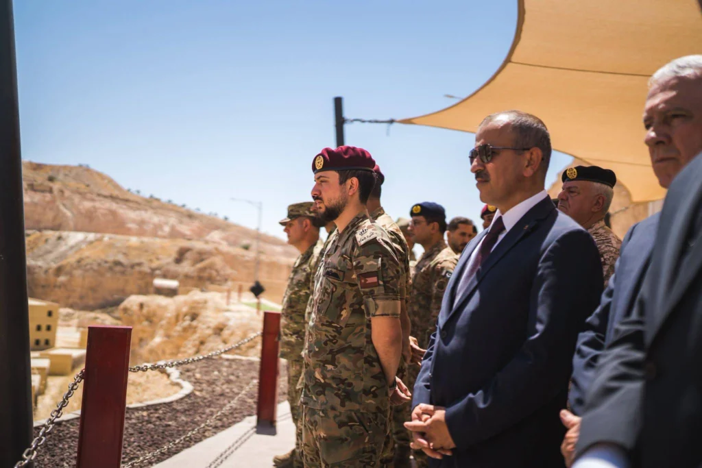 Jordanian Military Deterrence Collapses as Iran Strikes A formal outdoor photograph showing high-ranking Jordanian officials in dark suits and military officers in camouflage uniforms with maroon berets standing at attention under a canopy, overlooking a distant rugged outpost, symbolizing the defensive posture whose failure marks the collapse of Jordanian Military Deterrence against Iranian strikes.