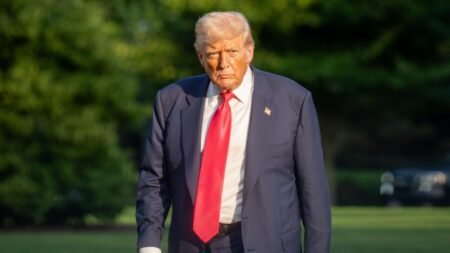 A photograph of a stern-looking President Donald Trump in a navy suit and red tie walking outdoors, his serious expression reflecting the mounting strategic frustration as the prolonged Iran War of Attrition defeats the US economic campaign strategy.