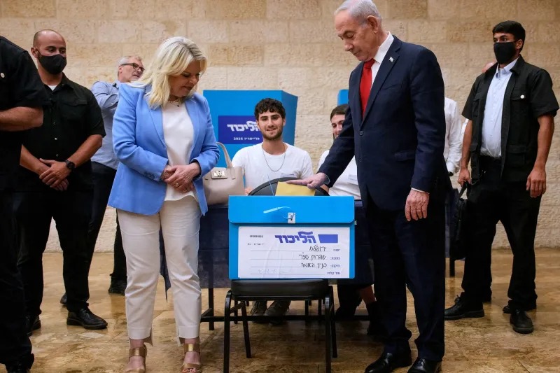 Israel Forever Wars: Can Eisenkot Break the Cycle? A photograph of Israeli Prime Minister Benjamin Netanyahu casting his ballot into a blue election box alongside his wife Sara Netanyahu, surrounded by security personnel, symbolizing the entrenched political status quo whose recurring conflicts perpetuate the Israel Forever Wars cycle that opposition leader Gadi Eisenkot seeks to break.