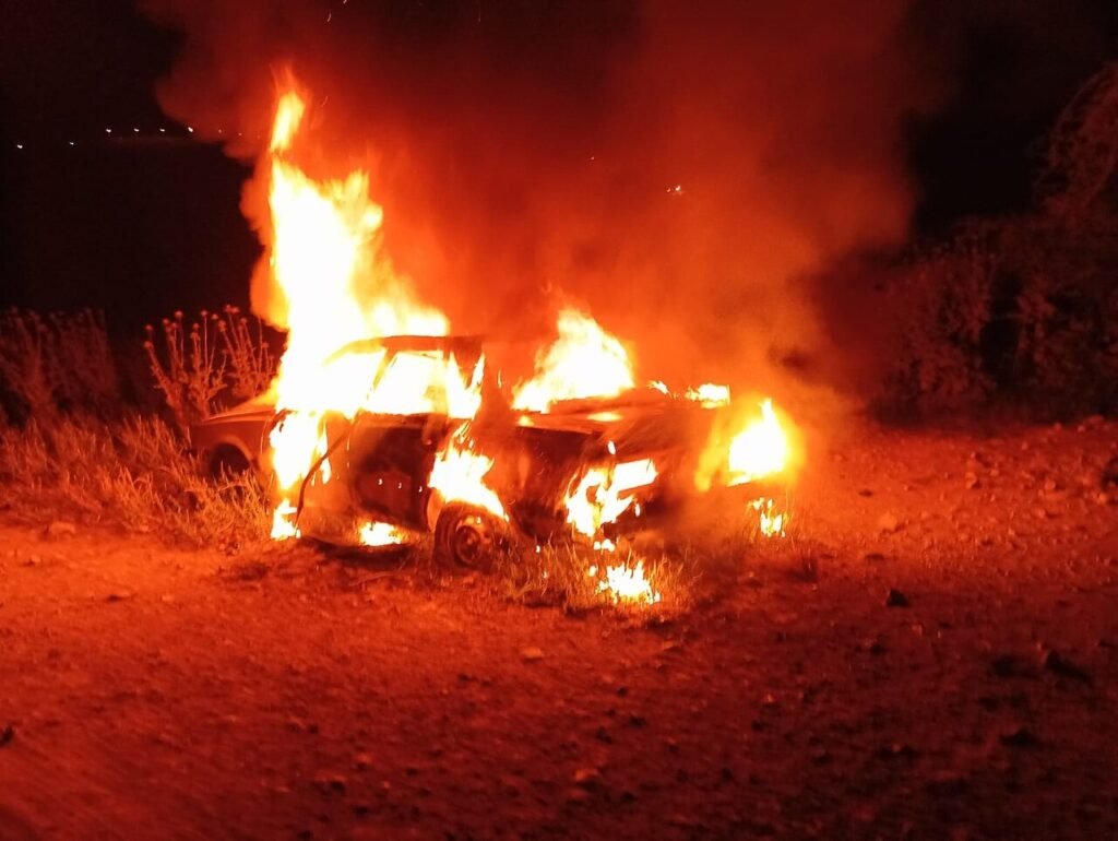 A burning car engulfed in heavy flames and smoke on a dark dirt road, symbolizing the Jewish Terrorism that a policy enforcement refuses to stop.