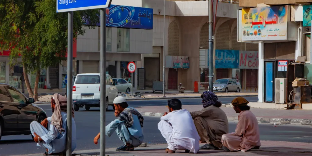 A street-level photograph showing several men in traditional Gulf attire sitting and crouching on a curb, waiting for day labor, against a backdrop of quiet storefronts and parked cars, symbolizing the stagnant employment market that continues to stall the Gulf Economic Recovery after six months of regional war.