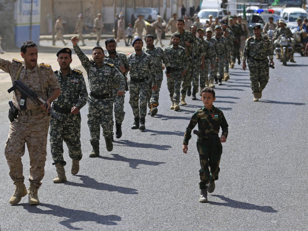Uniformed men and a young boy in camouflage march down a paved street, one man raising a fist and shouting, revealing desperation, not strength, in the Iran-Houthi Oil War.