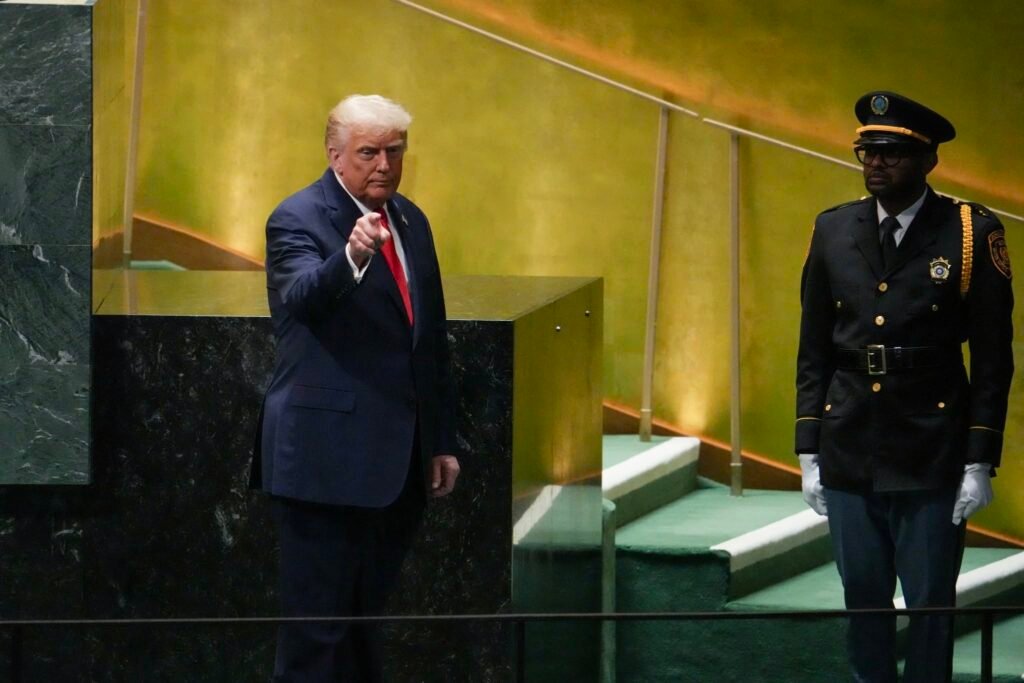 خطاب ترامب في الأمم المتحدة يضع عبء الاختيار على طهران Donald Trump in a dark blue suit and red tie points his finger forward while standing beside a dark marble podium at the United Nations, with a uniformed security officer in gold-trimmed dress uniform and white gloves standing behind him — the setting of a Trump UN Speech that puts the burden of choice on Tehran.