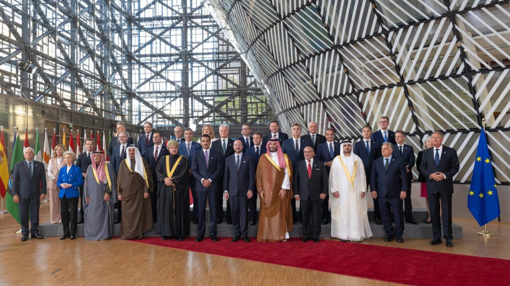 A formal group photograph of European and Gulf Arab leaders standing on a red carpet at a modern glass EU-GCC summit venue in Brussels, flanked by EU flags and member state flags, symbolizing the critical diplomatic reliance on Gulf energy partners that exposes the deepening European energy vulnerability as the Iran war drags on.