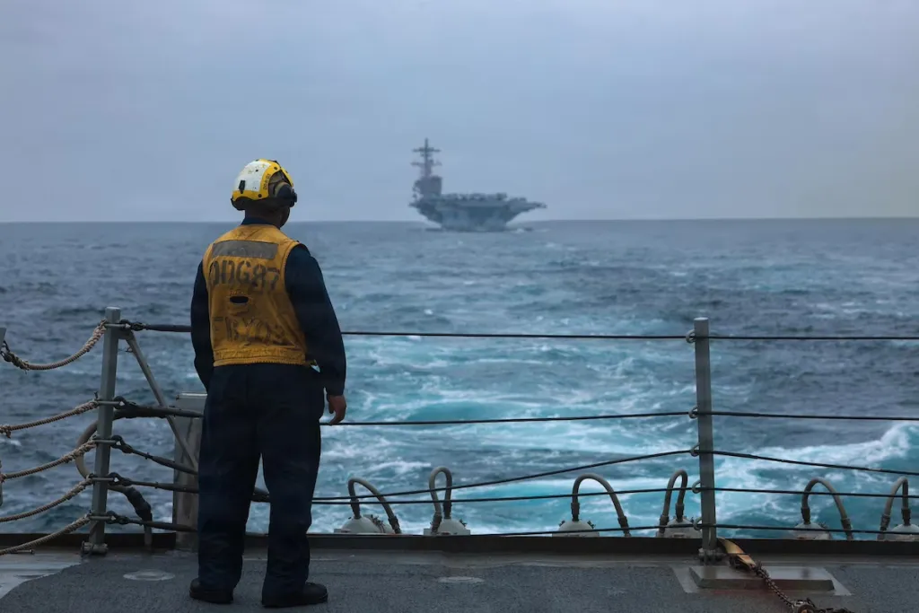 Iran survival is all Tehran needs to block US victory A sailor in a yellow vest on the deck of the USS Mason watches an aircraft carrier in rough, misty seas, depicting how Iran survival is all Tehran needs to block a US victory.