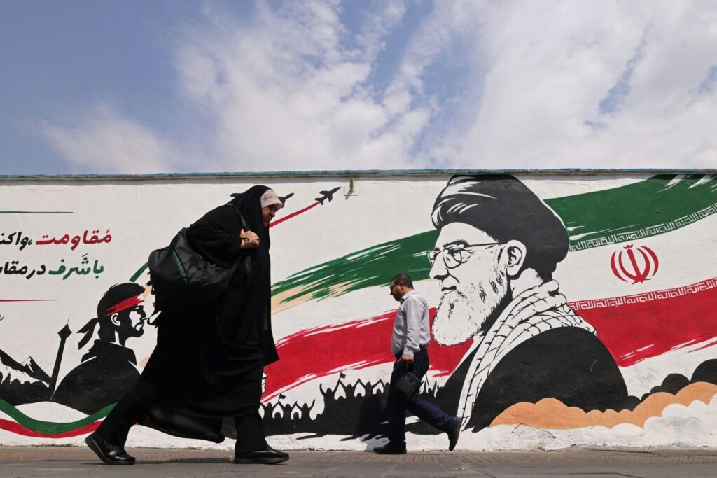Iran Oil Revenue, Not Ideology, Sustains the Regime A woman in a black chador and a man in a button-up shirt walk past a large wall mural of Ayatollah Khamenei painted in the colors of the Iranian flag, a scene of ordinary life passing beneath state ideology — a reminder that it is Iran Oil Revenue, not ideology, that sustains the regime.