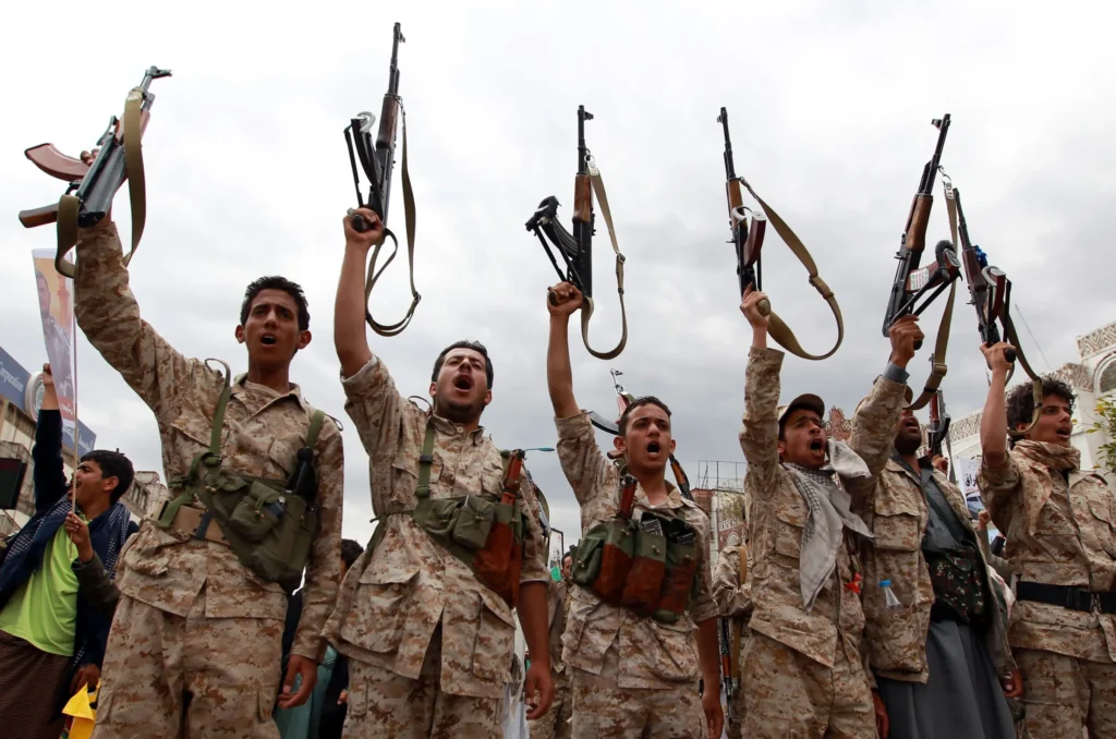 Uniformed Houthi fighters raising assault rifles and chanting during a rally, highlighting the ongoing military mobilization and the need for a political settlement in the Saudi-Houthi War.