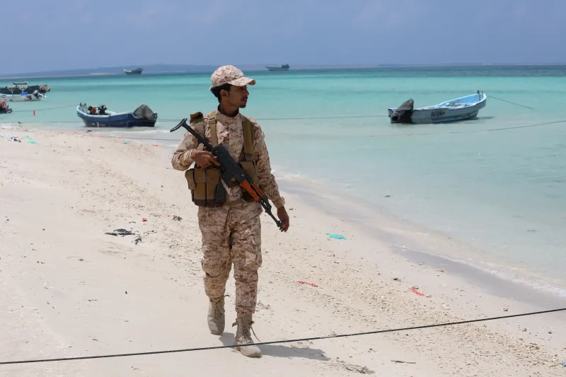 Saudi-UAE Rivalry Fuses Red Sea Theaters Into One Conflict A Yemeni soldier patrolling the coast near the Bab el-Mandeb Strait with small vessels in the background, highlighting coastal security dynamics influenced by the Saudi-UAE Rivalry.