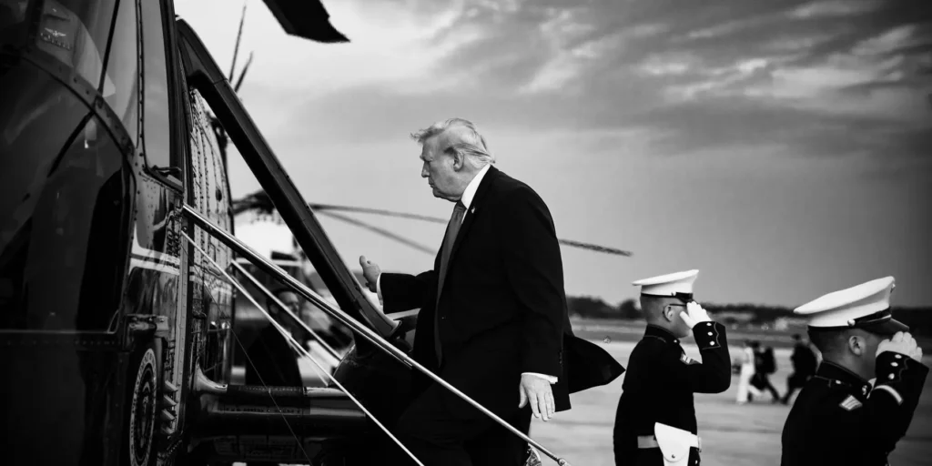 A black-and-white photo of Donald Trump boarding Marine One past saluting US Marines, illustrating the political and military tensions surrounding Iran Surrender Talk.