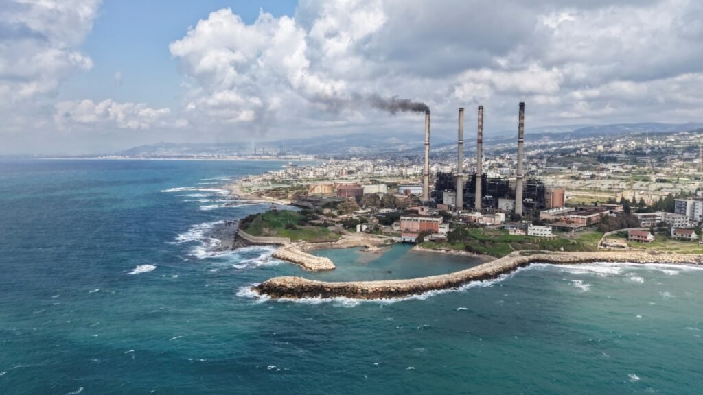 Aerial view of a coastal power plant in Baniyas with tall smokestacks emitting smoke, representing major energy infrastructure linked to Gulf Investment.