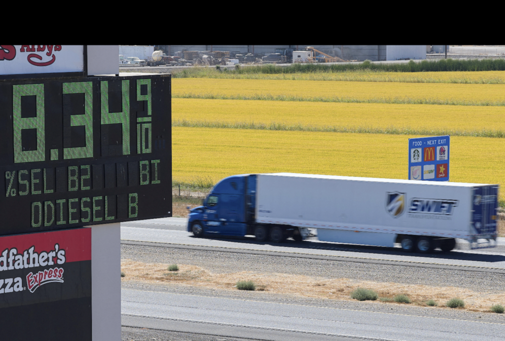 تكاليف حرب إيران: لماذا يدفع المزارعون ثمن إغلاق هرمز A digital gas station sign displaying high diesel prices beside a semi-truck driving past agricultural fields, illustrating how the Strait of Hormuz closure inflates fuel prices and Iran War Costs for farmers.