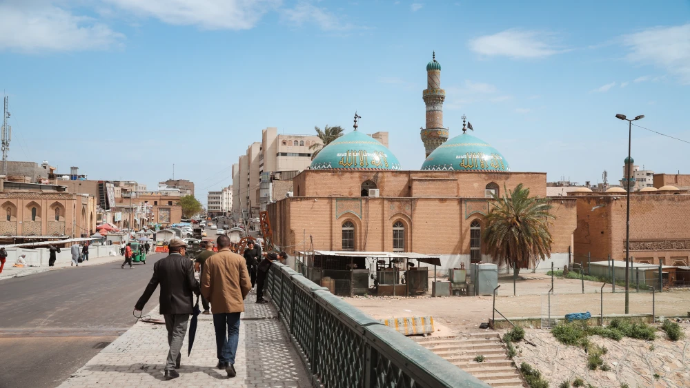 انهيار النظام الإيراني قد يؤدي إلى تقسيم العراق Pedestrians walking along a bridge overlooking an Iraqi city street with a traditional mosque featuring turquoise domes and a minaret, reflecting political uncertainty and the potential risk of Iraq Partition.