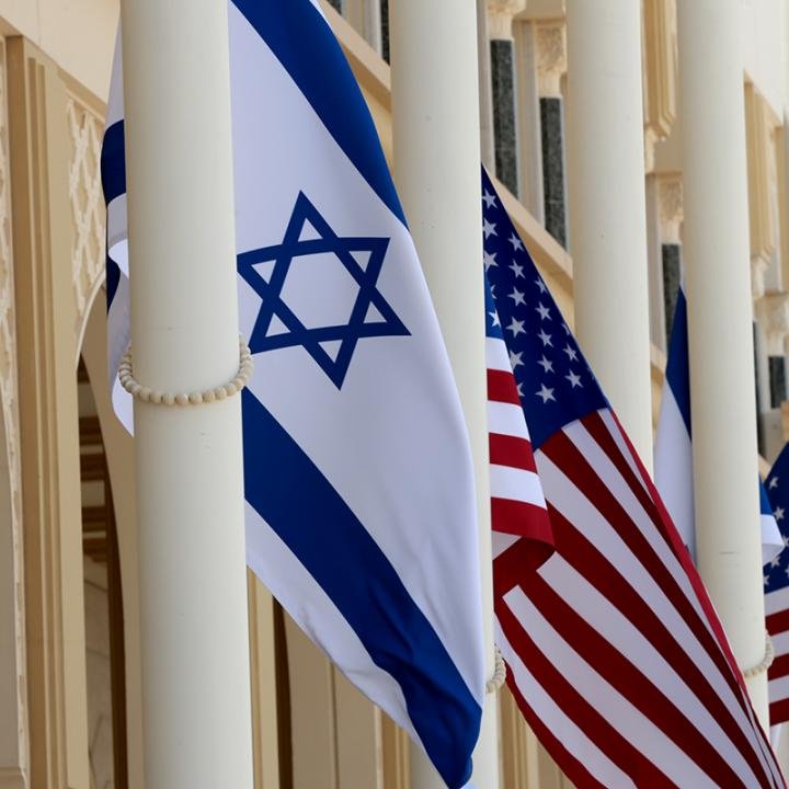 روابط ایالات متحده و اسرائیل در حال تضعیف است در حالی که نامزدها به آن توجه نمیکنند Israeli and American flags flying side-by-side on white pillars, illustrating the eroding US-Israel Relations as political candidates look away.