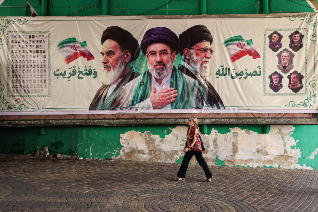 A woman walking past a large public banner in Tehran featuring Iranian supreme leaders, illustrating political resilience and leverage surrounding Hormuz tolls.