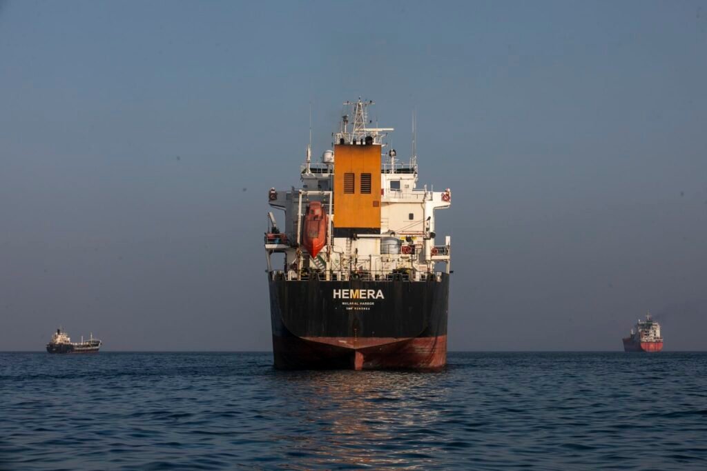 A large tanker named HEMERA sits anchored in the calm waters of the Strait of Hormuz near Iran's Larak Island, with two smaller vessels on the horizon — a scene showing the Geopolitical Chokepoints that let Iran turn cheap arms into power.