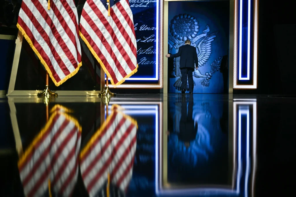 Donald Trump walking off stage toward a doorway emblazoned with the Presidential Seal flanked by American flags, illustrating political vulnerability ahead of the Midterm Elections.