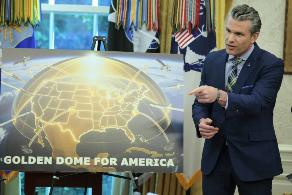 U.S. Secretary of War Pete Hegseth pointing at a "GOLDEN DOME FOR AMERICA" presentation board, illustrating the strategic missile defense plans for the American Golden Dome.
