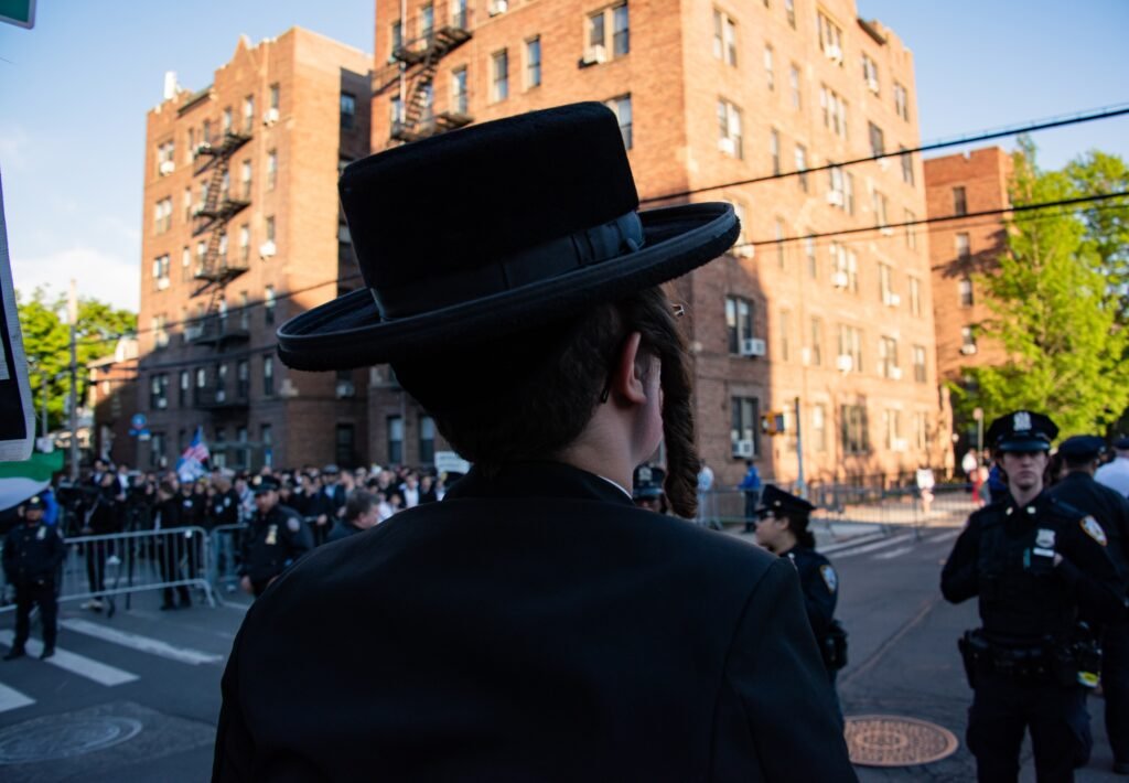 Diaspora Jews Now Stakeholders in Israel’s Policy Choices Rear view of an Orthodox Jewish man wearing traditional attire standing near a gathering with police officers, illustrating how Diaspora Jews engage with Israel's political decisions.