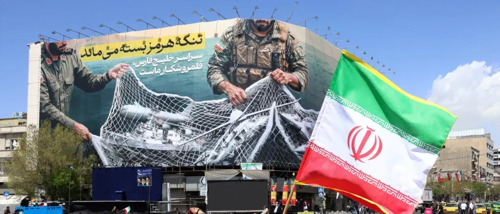 霍尔木兹海峡油轮战进入可持续僵局 A large propaganda billboard in Iran depicting military personnel holding a net over US naval ships and aircraft, with an Iranian flag in the foreground, reflecting escalation in the maritime Tanker war.
