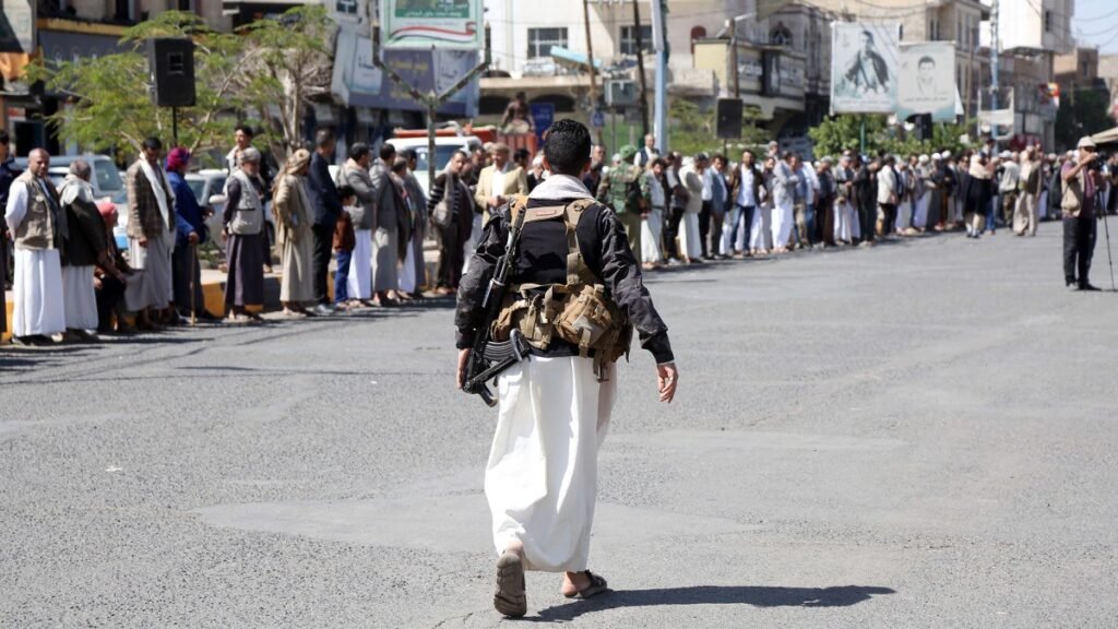 An armed Yemeni fighter in traditional white robes carrying a rifle down an empty street lined with spectators, illustrating political and military tensions in the Yemen war.