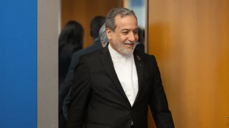 Iranian Foreign Minister Abbas Araghchi walking through a doorway with a slight smile, illustrating the internal political maneuvering and Iran Factionalism that leaves Mojtaba's authority in doubt.