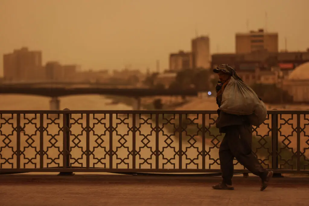 阿拉伯中产阶级在连环危机中崩解 A lone figure carrying a heavy sack across a bridge in a Middle Eastern city during a dusty, orange haze, illustrating economic strain impacting the Arab Middle Class.