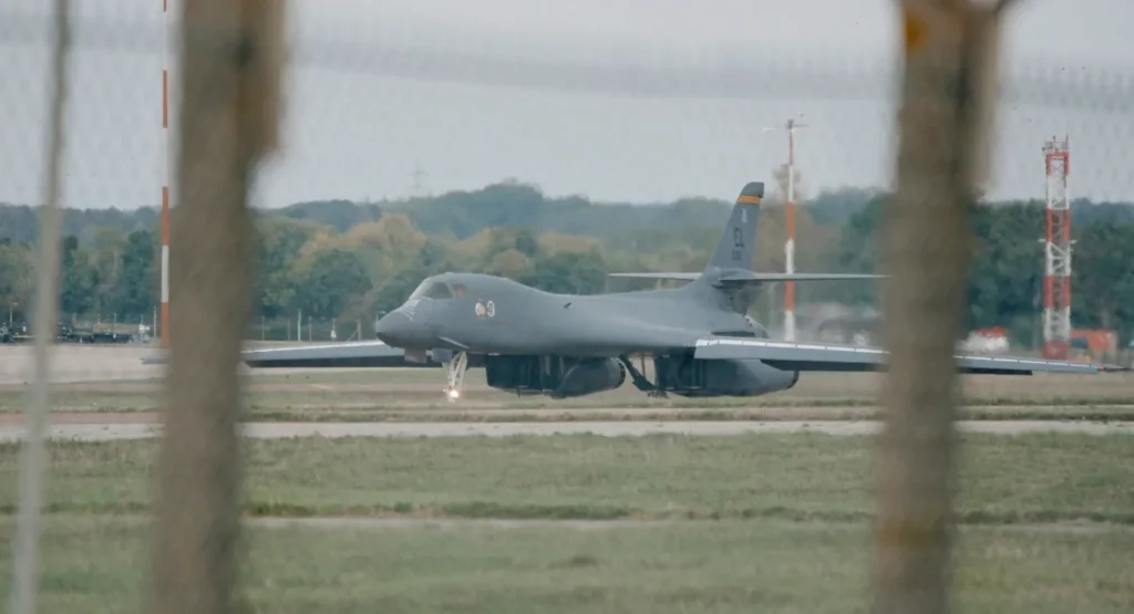 A U.S. B-1 Lancer bomber on the runway at RAF Fairford, viewed through a fence, illustrating the B-1 Bombers departing as Iran drones test U.S. defenses.