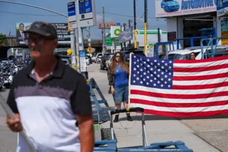 A woman holds a large American flag along a sunny Queens street as a motorcycle procession and a hearse carrying US Army Sergeant Angel Sarah Rampersad pass by, a scene of mourning that reflects the human cost of the Iran War Impact.