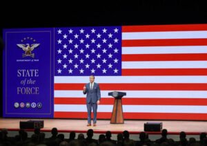 Pete Hegseth speaking before a large American flag at a State of the Force event, illustrating the Pentagon Overhaul that blurs reform and populism.
