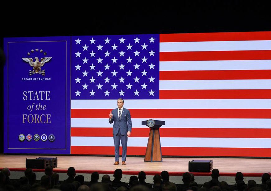 Pentagon Overhaul Blurs Line Between Reform and Populism Pete Hegseth speaking before a large American flag at a State of the Force event, illustrating the Pentagon Overhaul that blurs reform and populism.