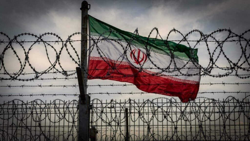 Iran-Backed Terrorism Exposes Limits of Sanctions Alone An Iranian flag fluttering behind a high fence wrapped with razor barbed wire under a cloudy sky, symbolizing international sanctions enforcement and efforts to counter Iran-Backed Terrorism.