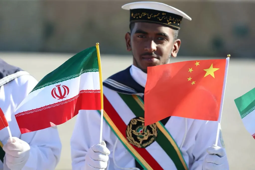 An Iranian sailor holding small Iranian and Chinese flags during a joint ceremony, demonstrating how broader US-China Rivalry shapes global sanctions enforcement and geopolitical alignments.