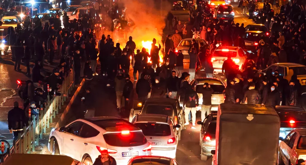 Iran Protests: Every Massacre Only Feeds the Next Uprising A large crowd gathered in traffic-congested city streets around a street fire during night-time Iran Protests, illustrating how state crackdowns ignite recurring public uprisings.