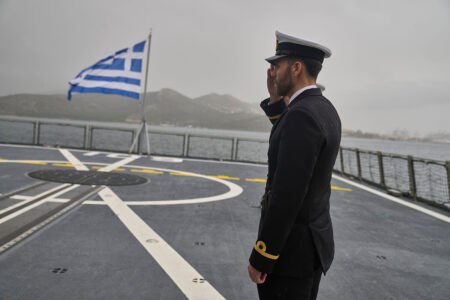 A Greek naval officer salutes on a ship deck with the Greek flag, representing European Military Power that Iran fears.